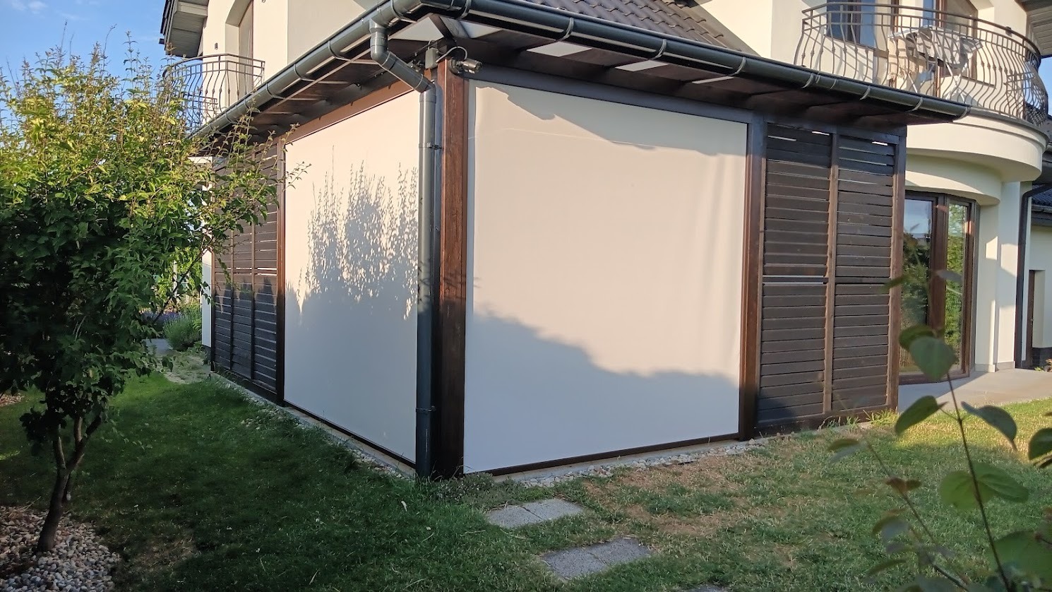 Zadaszony taras z beżowymi roletami bocznymi, drewnianą konstrukcją i elementami nowoczesnej architektury domu w tle. Widoczny fragment ogrodu z zielenią.