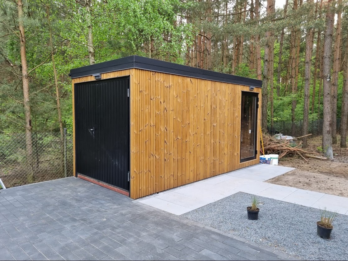 Nowoczesna, drewniana altana z czarnymi elementami, otoczona lasem, z wybrukowanym podjazdem i dekoracyjnymi roślinami w donicach. Minimalistyczny design.