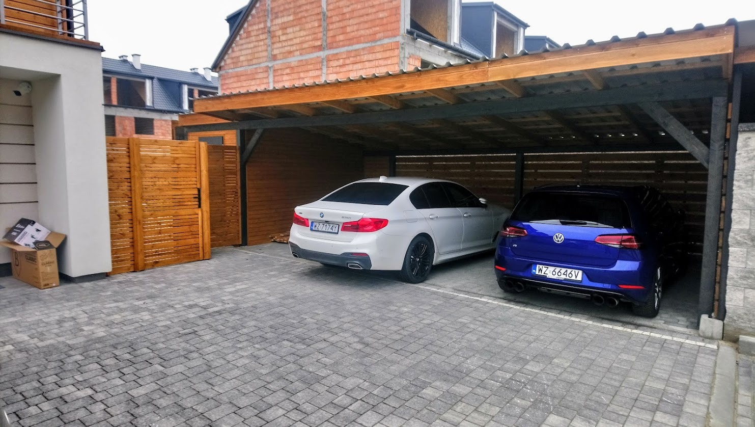 Zadaszone miejsce parkingowe z dwoma samochodami: białym BMW i niebieskim VW Golfem. Konstrukcja drewniana z elementami metalowymi. Kostka brukowa na podjeździe.