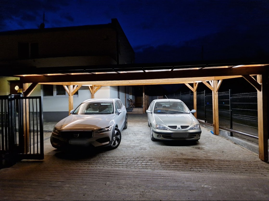 Dwa auta pod drewnianą, oświetloną pergolą z czarnym, metalowym dachem na posesji z kostki brukowej. Nocne ujęcie z widocznym budynkiem w tle.
