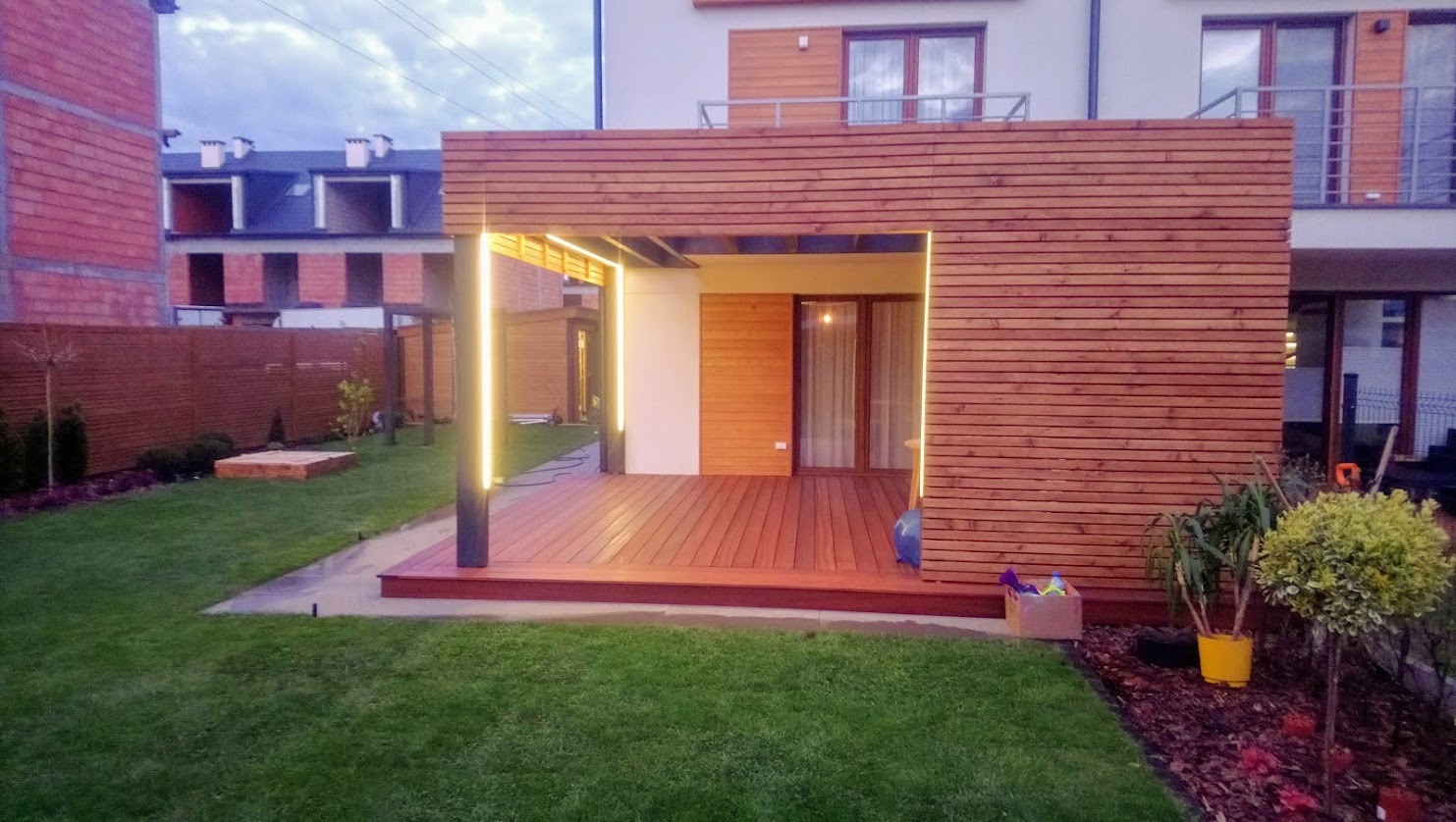 Nowoczesna, drewniana pergola z oświetleniem LED i tarasem z desek na trawniku. Widok na budynek mieszkalny w tle. Minimalistyczny design.