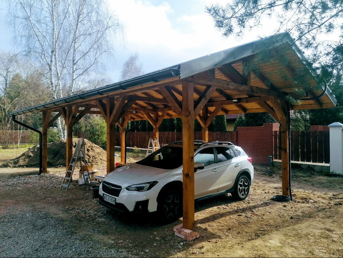 Drewniana wiata garażowa z ciemnym dachem i orynnowaniem, chroniąca biały samochód. W tle drzewa i częściowo widoczny mur z cegły.