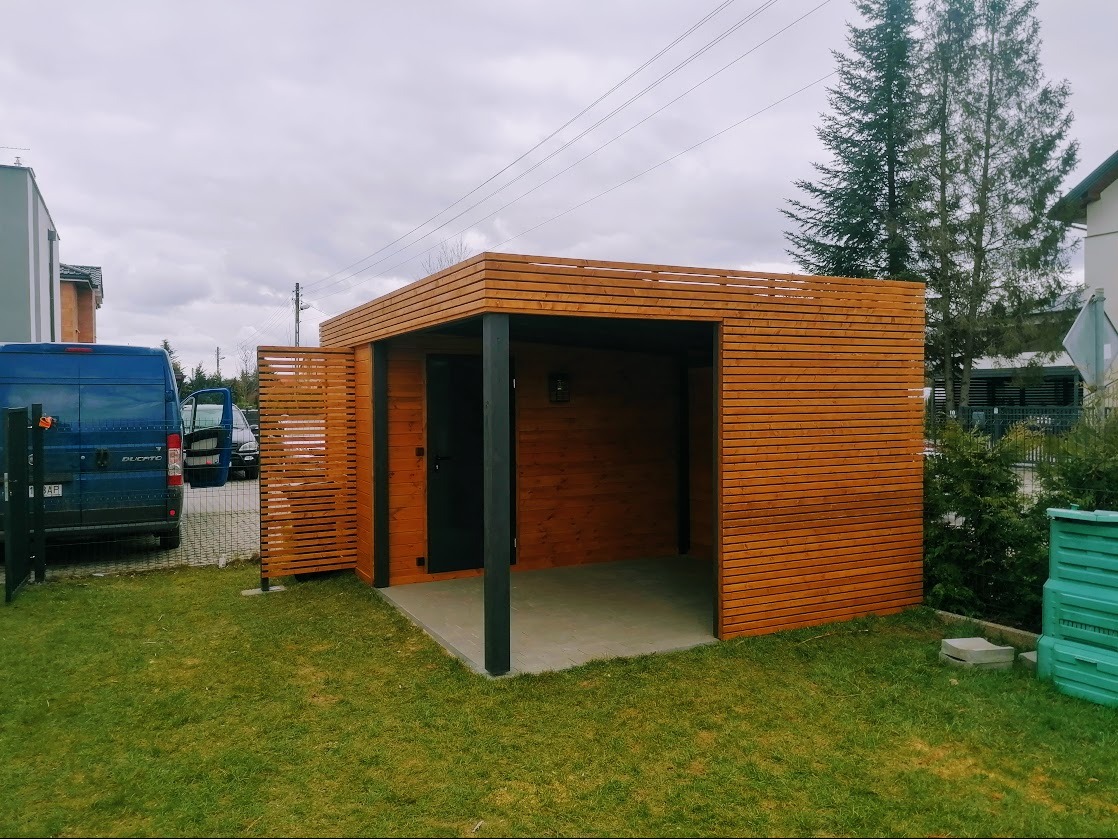 Nowoczesna pergola z drewna w Feliksowie. Konstrukcja z poziomych desek, czarne słupy, szara podłoga. Na trawniku, obok zaparkowany bus.