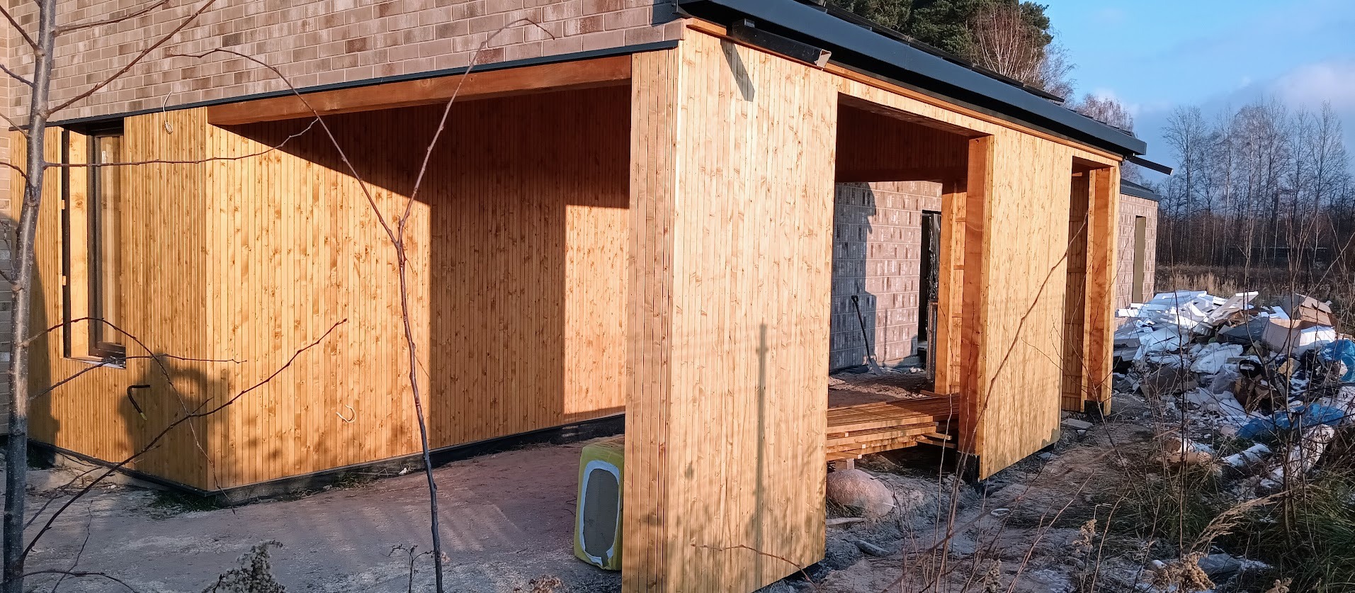 Surowa drewniana konstrukcja z widocznymi elementami budynku w tle, ukazana w jasnym świetle słonecznym, z widocznymi śladami budowy.