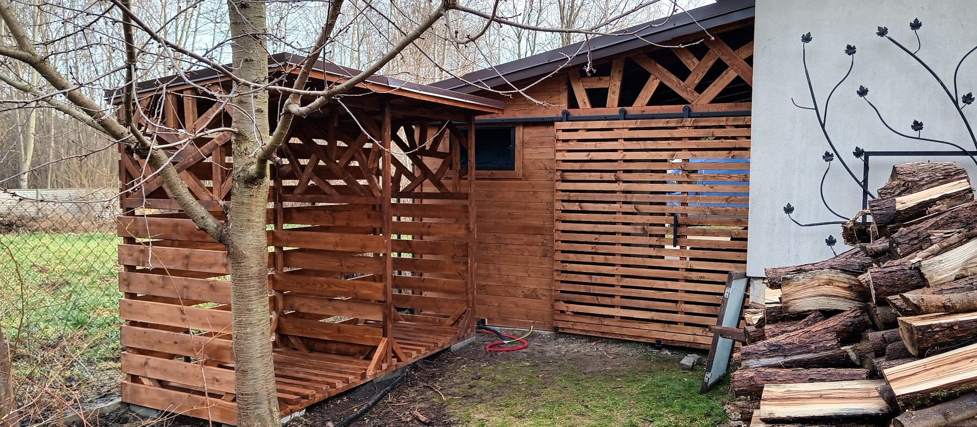 Drewniana wiata na drewno z przesuwanymi drzwiami i dekoracją ścienną z motywem roślinnym. Konstrukcja z bejcowanego drewna na zewnątrz.