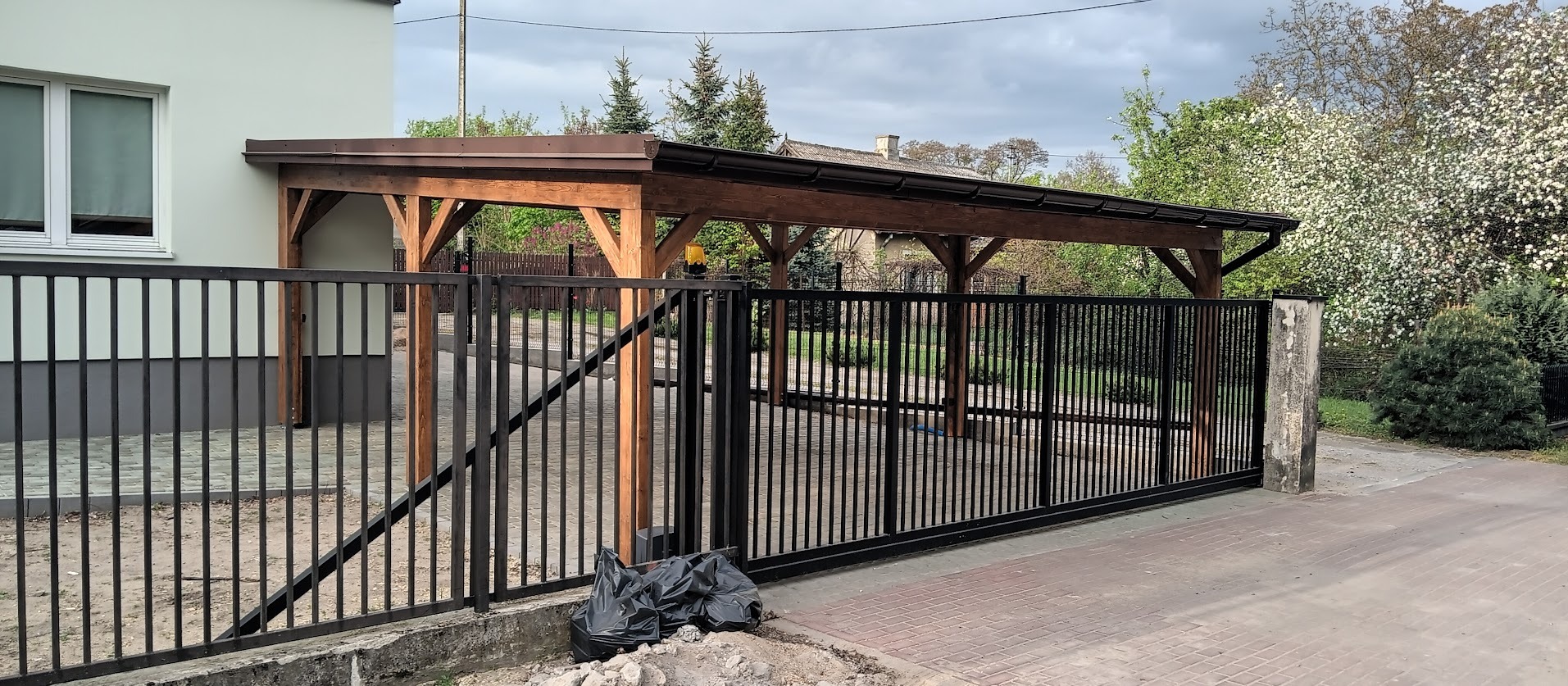 Drewniana wiata samochodowa z brązowym dachem, widoczna za czarnym, metalowym ogrodzeniem z bramą przesuwną. W tle zieleń drzew i krzewów, pochmurne niebo.