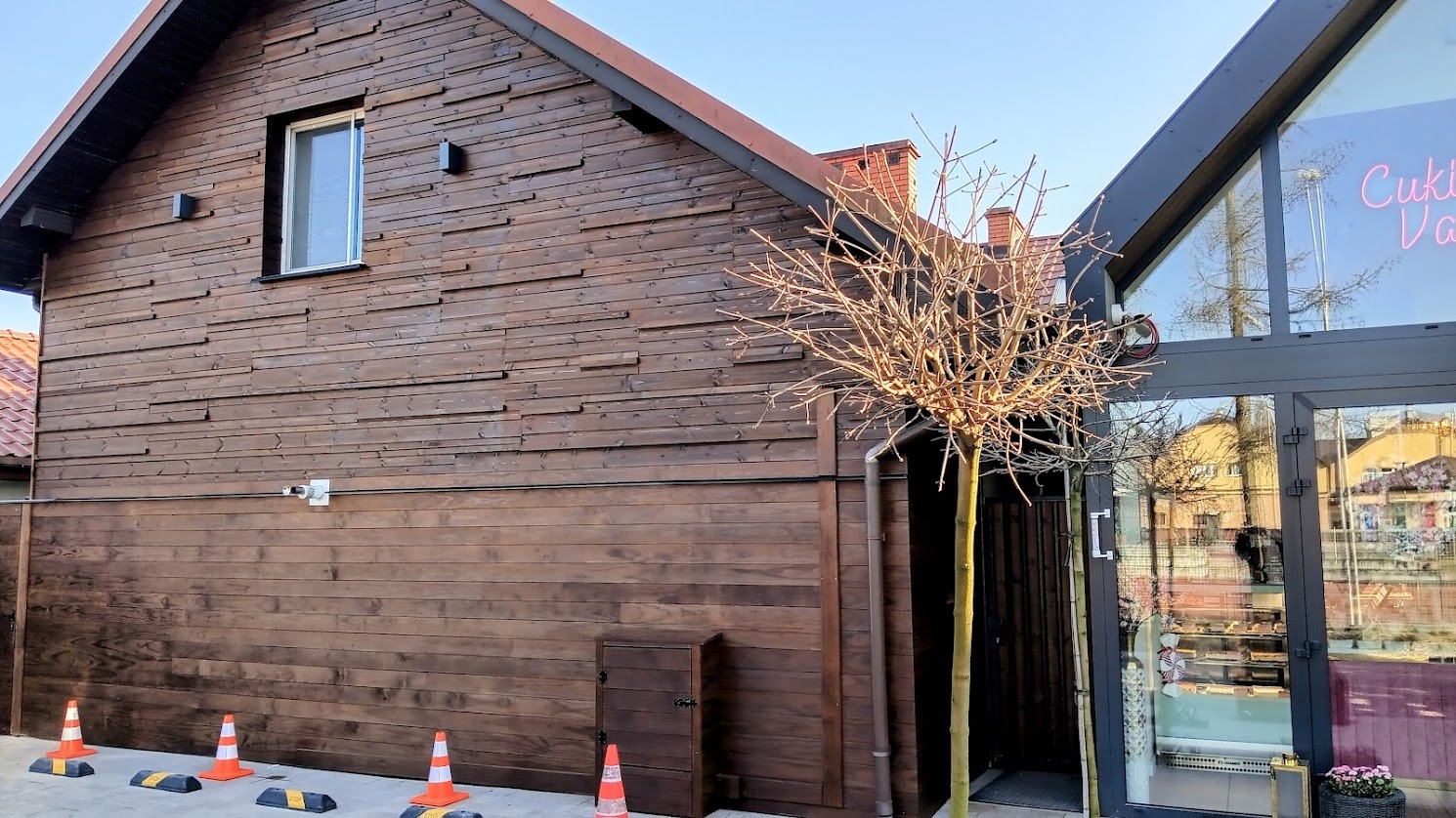 Elewacja budynku obita ciemnym drewnem, z widocznymi poziomymi deskami. Małe okno na górze, rynna i małe drzwi na dole. Pomarańczowe pachołki na chodniku.