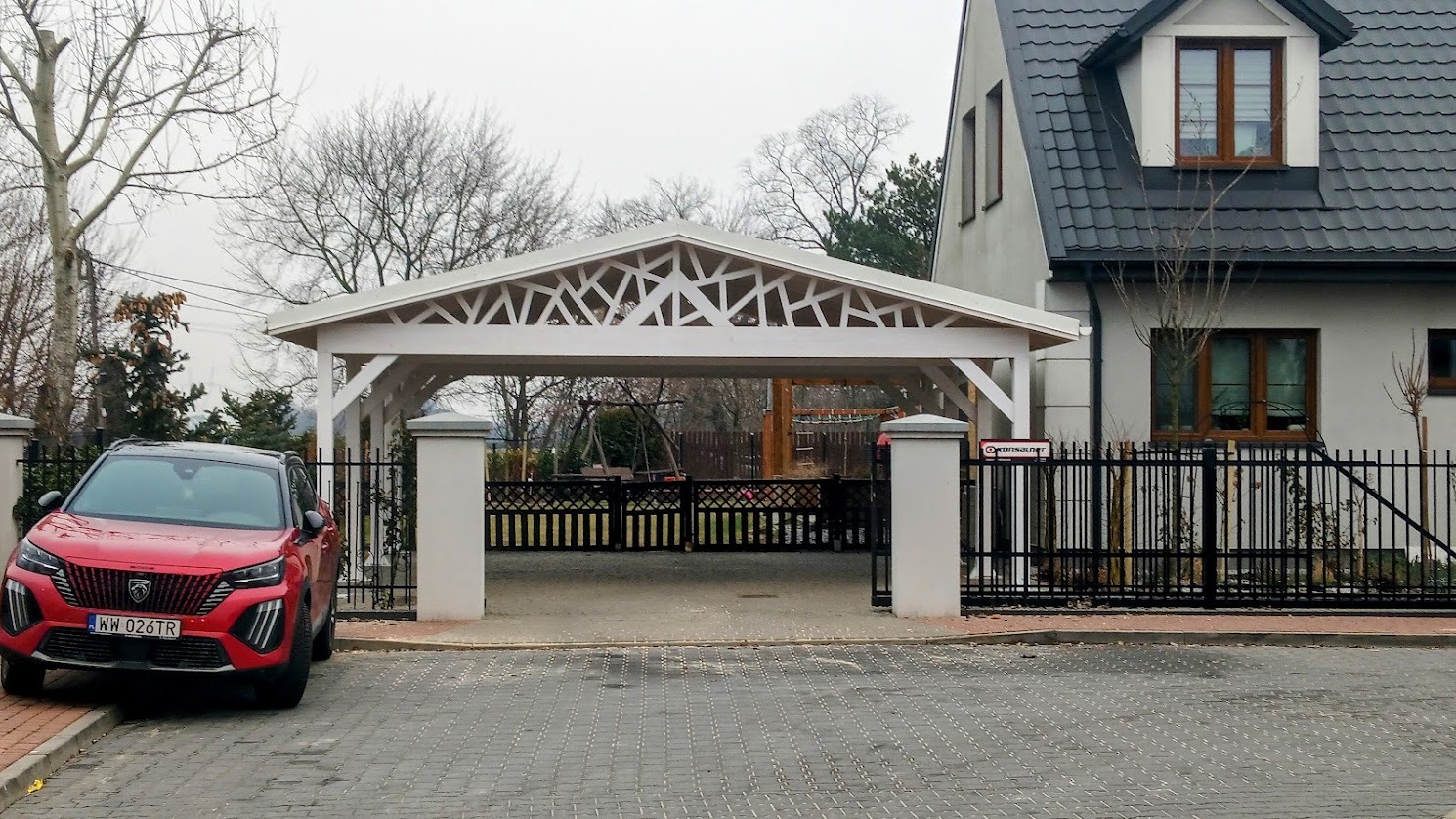 Biała wiata garażowa z dekoracyjnym ażurem, czerwony samochód na kostce brukowej, nowoczesny dom z ciemnym dachem i czarnym ogrodzeniem w tle.