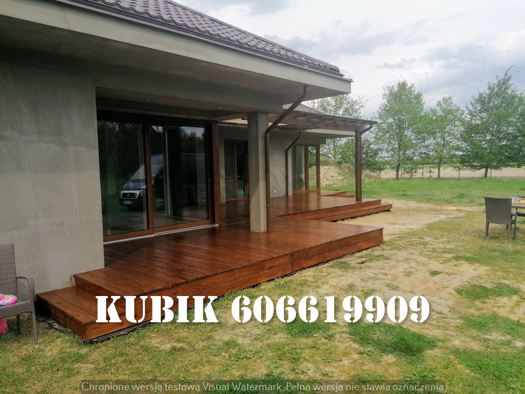 Nowoczesny taras drewniany zadaszony pergolą, brązowe deski, widok z zewnątrz, szklane drzwi przesuwne, trawnik. Widoczny numer telefonu na desce.