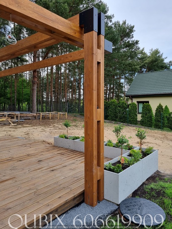 Drewniana pergola z czarnymi łącznikami, betonowe donice z roślinami, deski tarasowe. Widok na ogród z domem w tle. Ujęcie z dołu, podkreślające konstrukcję.