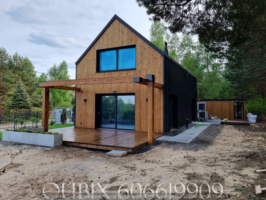 Nowoczesny dom z drewnianą elewacją i tarasem, połączony z pergolą. Widoczne czarne panele, okna z niebieską folią, otoczenie leśne, świeżo zagospodarowany teren.