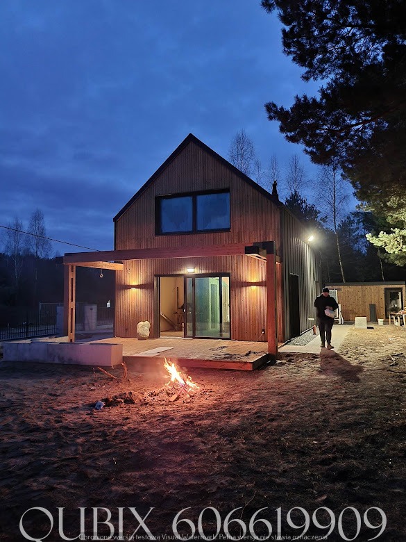 Nowoczesny dom z drewnianą elewacją i pergolą oświetloną lampami, ognisko na pierwszym planie, osoba w tle. Ujęcie wieczorne.