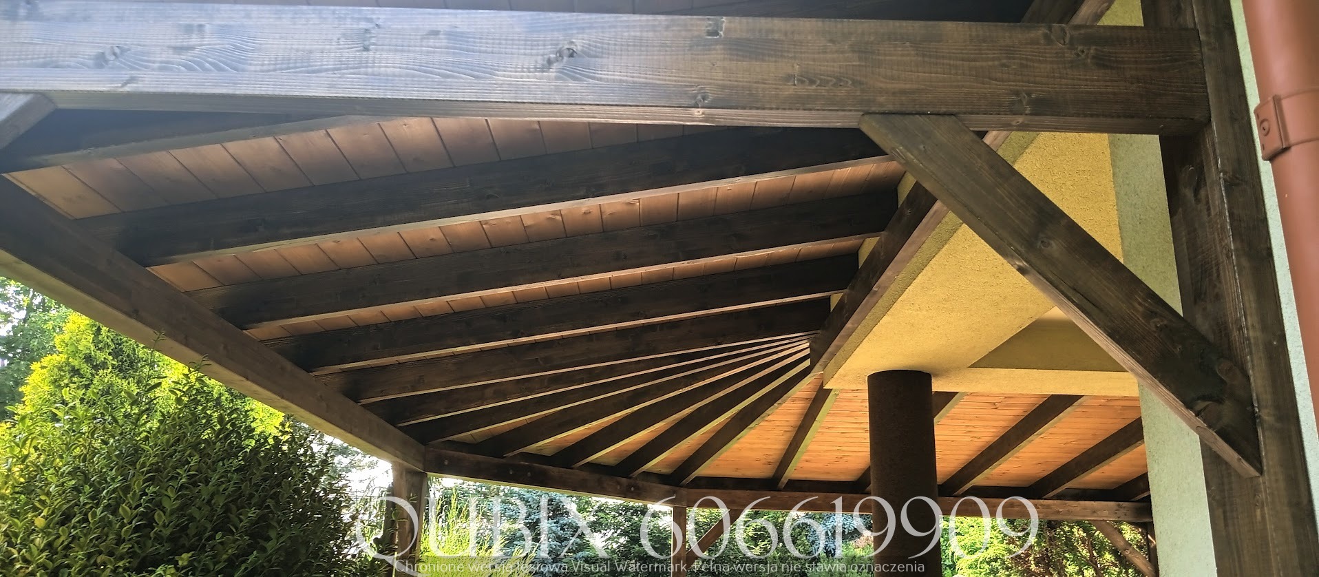 Drewniana konstrukcja z zadaszeniem, widok od dołu. Ciemne, bejcowane belki tworzą geometryczny wzór na tle zieleni. Elementy budynku z widoczną teksturą.