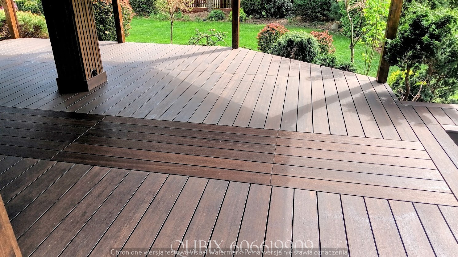 Zadaszony taras z desek kompozytowych w odcieniach brązu. Widoczne słupy konstrukcyjne i zieleń ogrodu w tle, tworzące spójną przestrzeń relaksu na świeżym powietrzu.