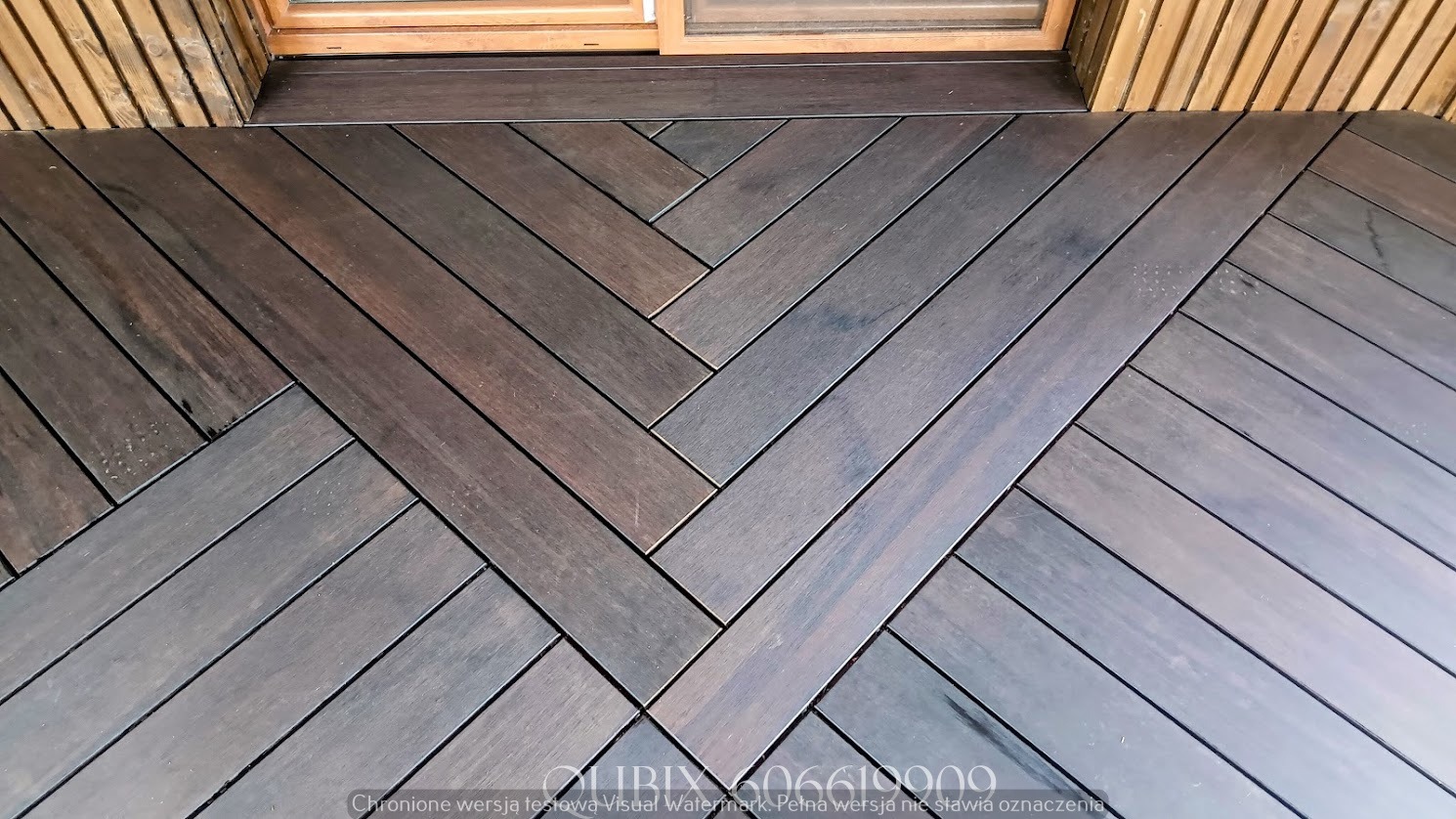 Ciemny taras drewniany ułożony we wzór jodełki, widok z góry. Deski o wyraźnym usłojeniu, z widocznym oknem i fragmentem drewnianej elewacji w tle.