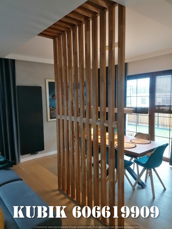 Drewniana ścianka działowa z pionowych listew oddziela salon od jadalni. Widoczny stół z krzesłami i okno z roletami w tle. Ujęcie z niskiego kąta.