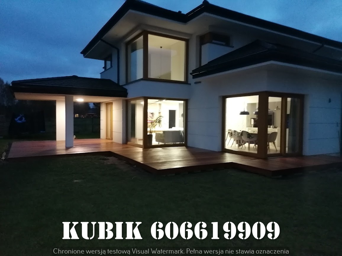 Nowoczesny dom z drewnianym tarasem oświetlonym wieczornym światłem, widok z ogrodu. Duże okna z drewnianymi ramami. Widoczny fragment trawnika.