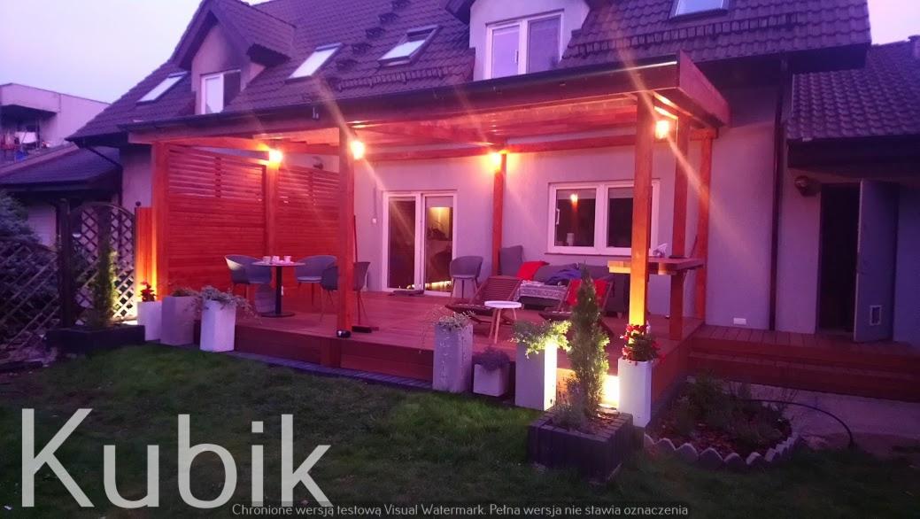 Oświetlony taras z drewna i pergola przy domu jednorodzinnym, z drewnianą ścianką działową, meblami ogrodowymi i dekoracyjnymi donicami z roślinami, sfotografowany o zmierzchu.