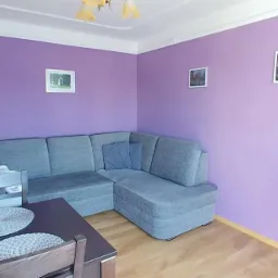 Pokój z fioletowymi ścianami, szarą narożną sofą, drewnianym stołem i krzesłami oraz dekoracjami ściennymi.