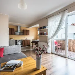 Jasne wnętrze mieszkania z otwartą kuchnią, balkonem i drewnianą podłogą; na stole wazon z kwiatami i magazyn.