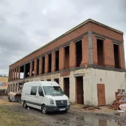 Dwukondygnacyjny budynek w trakcie murowania ścian z czerwonej cegły, z widocznymi otworami okiennymi i drzwiami, na pierwszym planie biały samochód dostawczy z przyczepką na błotnistym terenie.