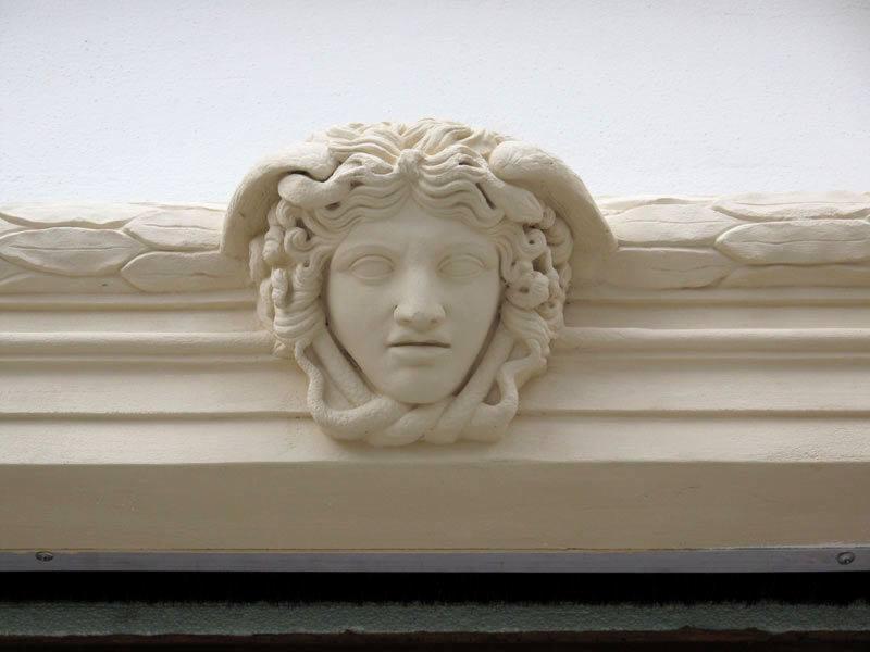 Dekoracyjny, kremowy gipsowy ornament przedstawiający głowę Meduzy z wężami zamiast włosów, umieszczony nad listwą przypodłogową w pomieszczeniu.