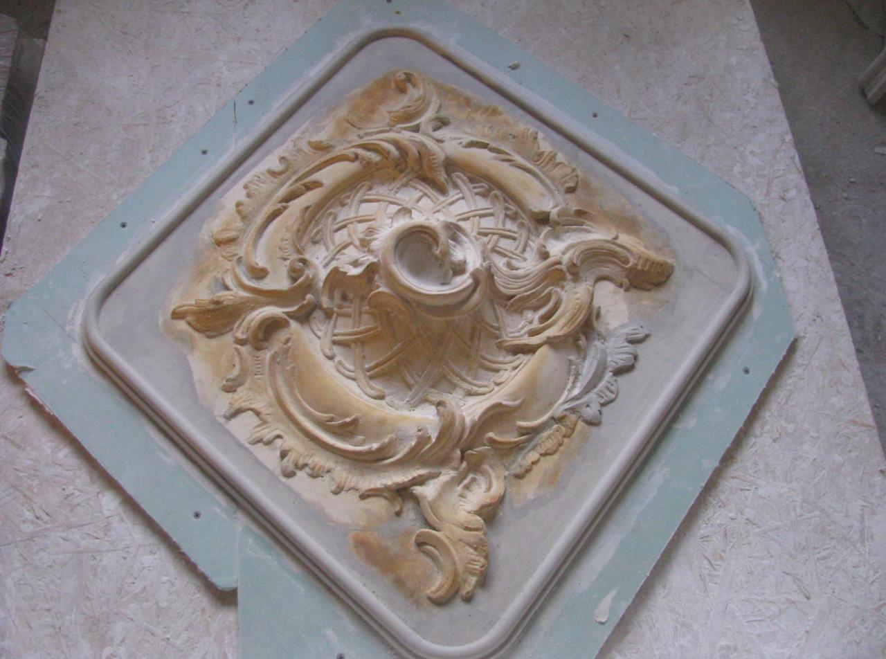 Gipsowy element dekoracyjny w formie rozety z ornamentami, umieszczony na płycie gipsowej, prawdopodobnie w trakcie tworzenia lub obróbki.