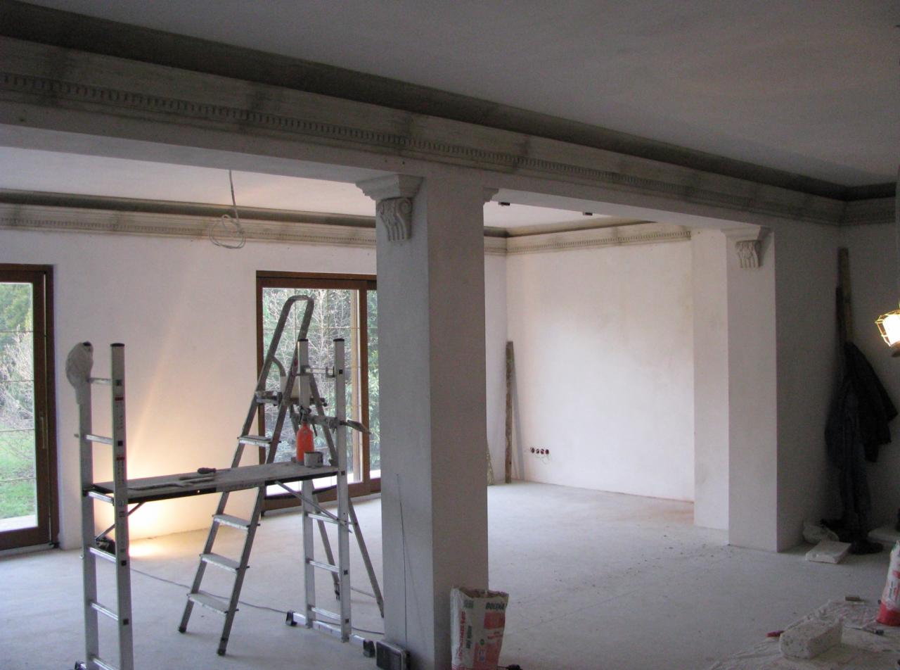 Wnętrze w trakcie prac wykończeniowych z kolumnami i sztukaterią, widoczne rusztowanie, gładkie, białe ściany, surowy beton na podłodze, okna z widokiem na zieleń.