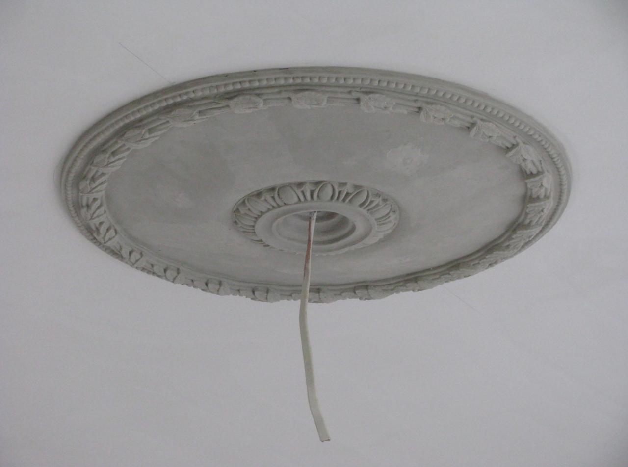 Niedokończona, gipsowa rozeta sufitowa z dekoracyjnym ornamentem, z otworu wystaje kabel elektryczny.