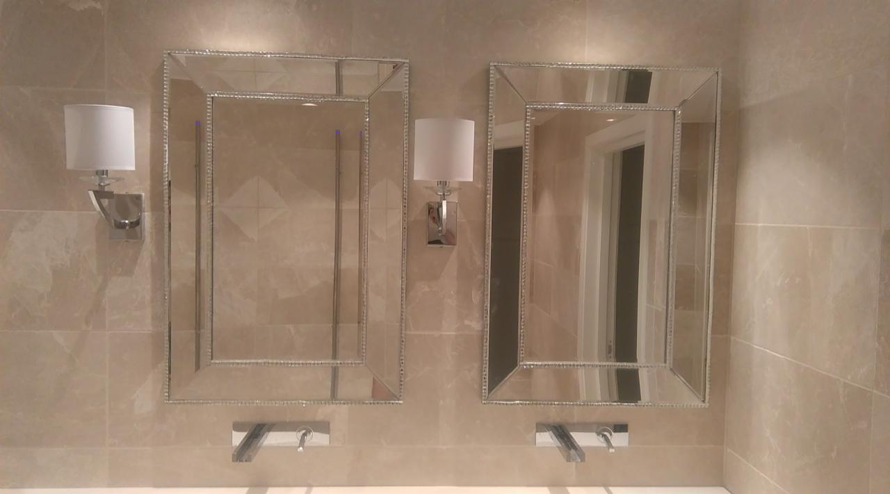 Dwa prostokątne lustra z dekoracyjną ramą, umieszczone nad umywalkami z chromowanymi kranami, oświetlone kinkietami z białymi abażurami, na tle beżowych płytek imitujących marmur.
