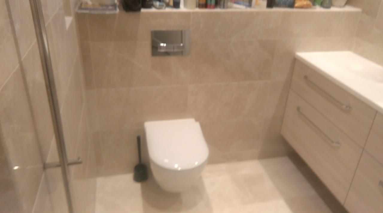 Nowoczesna łazienka w beżowych płytkach z podwieszaną toaletą, szafką pod umywalką z szufladami i chromowanym przyciskiem spłukującym. Widoczny fragment kabiny prysznicowej i szczotka do WC.