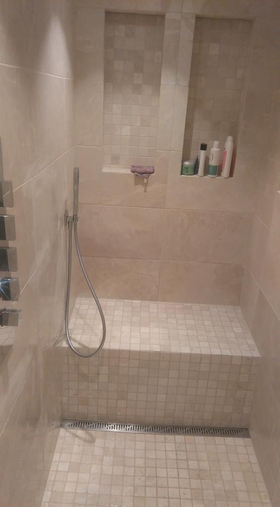 Beżowa kabina prysznicowa z siedziskiem wyłożona mozaiką, z odpływem liniowym i wnękami na kosmetyki.