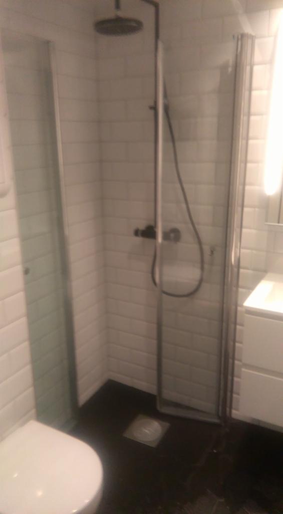 Nowoczesna kabina prysznicowa z deszczownicą i szklanymi drzwiami, wykończona białymi, cegiełkowymi płytkami i czarną podłogą z odpływem liniowym.