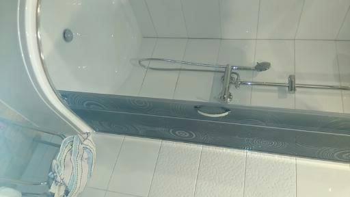 Biała wanna z szarą obudową z dekoracyjnym wzorem w łazience wyłożonej białymi kafelkami w Berlinie, widoczny chromowany zestaw prysznicowy.