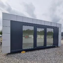 Gotowy modułowy budynek z trzema dużymi oknami i drzwiami, ustawiony na żółtych podkładkach na żwirowym placu, z widocznym w tle wózkiem widłowym i stosem płyt warstwowych.