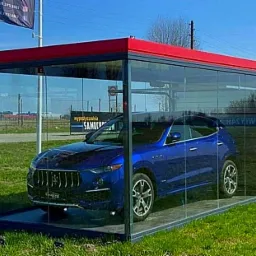 Niebieski samochód osobowy w przeszklonej, stalowo-czerwonej konstrukcji na trawniku, z widocznymi plakatami reklamowymi na ścianach.