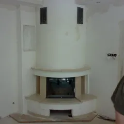 Wnętrze z niedokończonym kominkiem z obudową z jasnego kamienia, widoczne ślady prac wykończeniowych na ścianach.