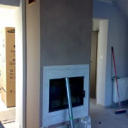 Nowoczesny portal kominkowy z szarą ścianą, w trakcie montażu, widoczna poziomica i materiały wykończeniowe na podłodze.