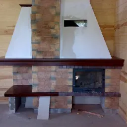 Kominek z obudową z kamienia dekoracyjnego, z drewnianym blatem i półką, w trakcie wykańczania, widoczne elementy konstrukcyjne i surowe drewno na ścianach.