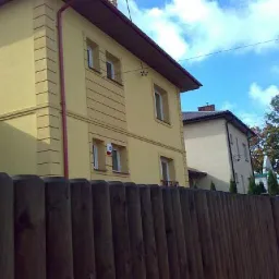 Żółta dwupiętrowa elewacja domu z dekoracyjnymi elementami, widoczna zza drewnianego płotu, z fragmentem drugiego budynku w tle.