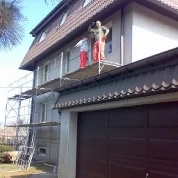 Pracownicy na rusztowaniu przy białej elewacji domu z brązowym dachem, jeden z nich w pomarańczowych spodniach opiera się o ścianę