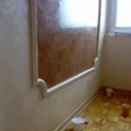 Ściana wewnętrzna wykończona dekoracyjnym tynkiem trawertyn, z widocznymi detalami imitującymi naturalny kamień, w pomieszczeniu w trakcie prac wykończeniowych, zabezpieczona folią podłoga.