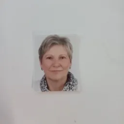 Zdjęcie portretowe kobiety z krótkimi siwymi włosami, umieszczone na białej ścianie z widocznym cieniem i drobnymi zabrudzeniami.