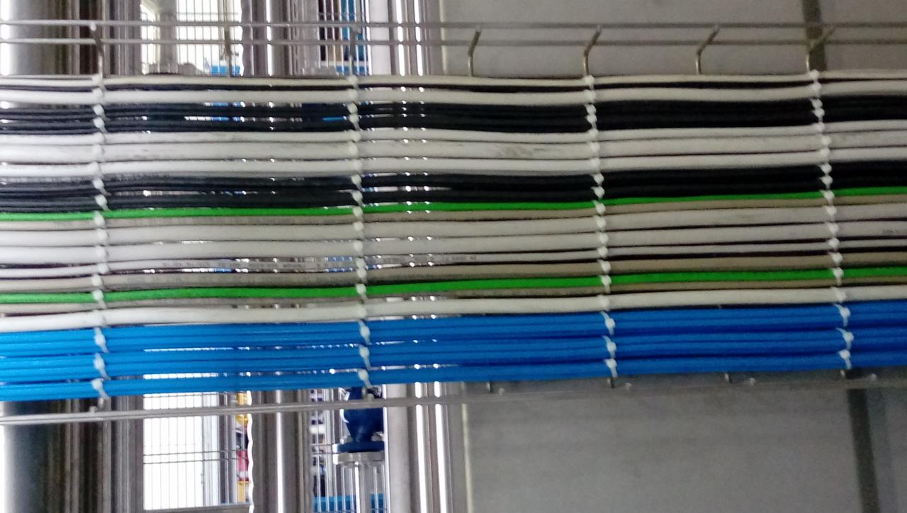 Rzędy uporządkowanych przewodów elektrycznych i rur wentylacyjnych w kolorach: białym, czarnym, zielonym i niebieskim, przymocowanych do metalowej konstrukcji za pomocą białych opasek zaciskowych.