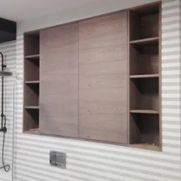 Drewniana szafa wnękowa z przesuwnymi drzwiami i otwartymi półkami w łazience z poziomymi pasami na ścianach, obok kabiny prysznicowej z czarną armaturą.