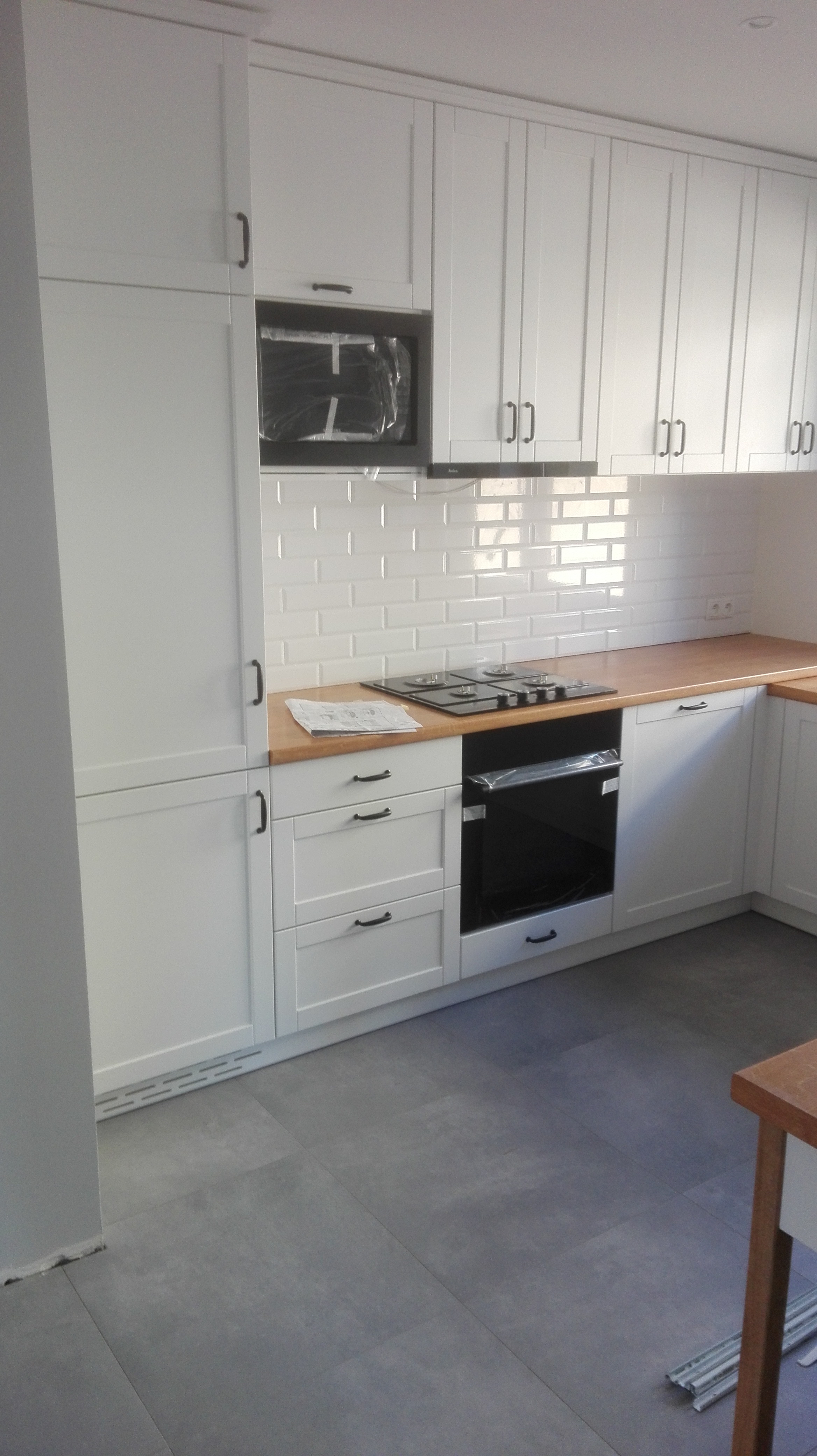 Biała zabudowa kuchenna z drewnianym blatem i piekarnikiem w folii ochronnej, widok z niskiego kąta.