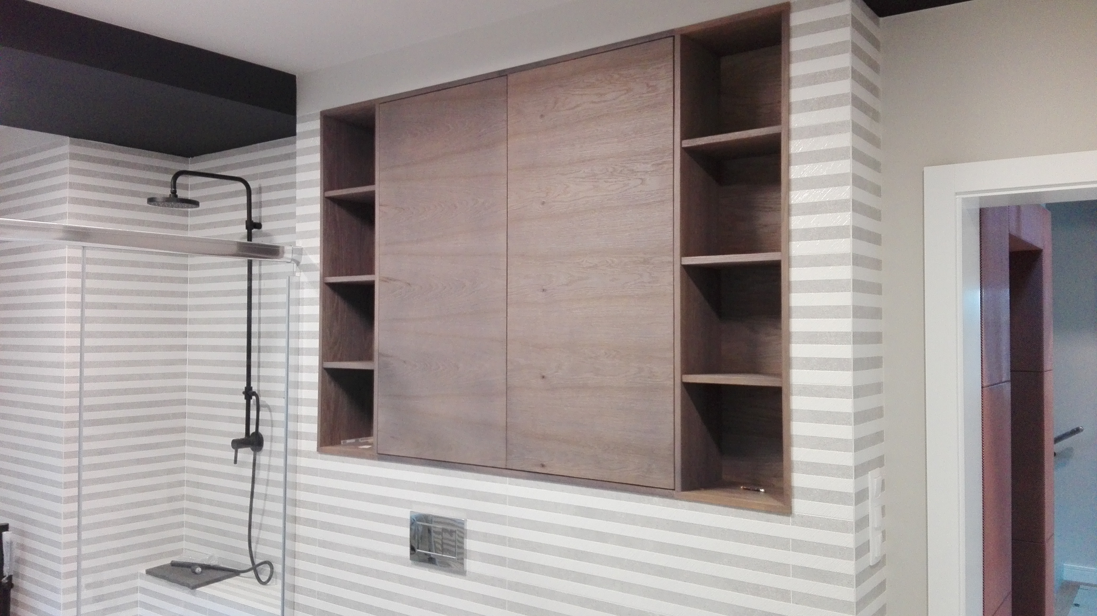 Drewniana szafa wnękowa z przesuwnymi drzwiami i otwartymi półkami w łazience z poziomymi pasami na ścianach, obok kabiny prysznicowej z czarną armaturą.