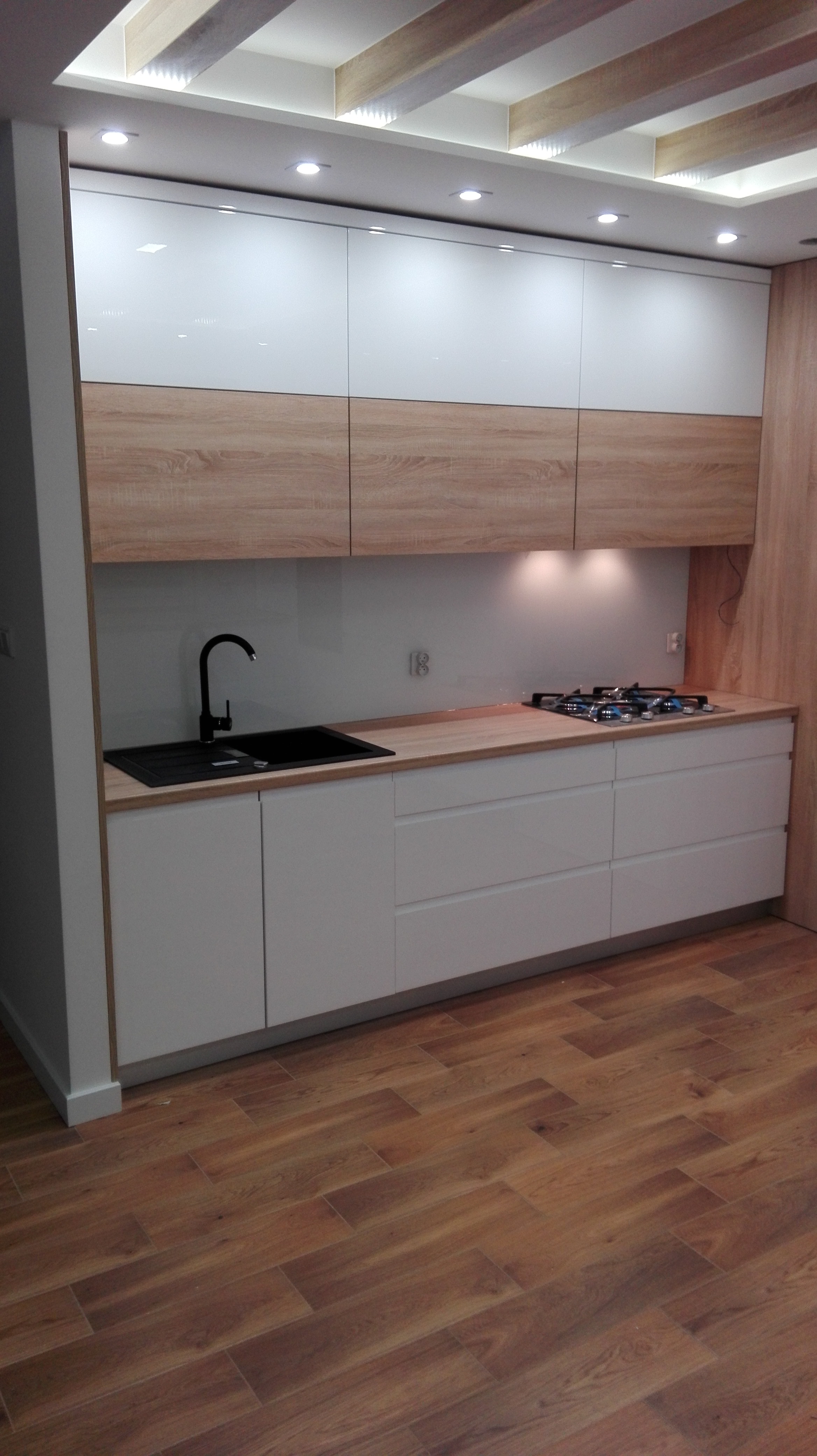 Nowoczesna kuchnia z białymi szafkami dolnymi i górnymi z frontami w kolorze jasnego drewna, czarną armaturą i płytą gazową, oświetlona punktowymi lampami w suficie.