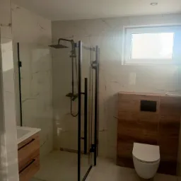 Nowoczesna łazienka z kabiną prysznicową typu walk-in z czarnymi profilami, umywalką z szafką z drewnopodobnym frontem i podwieszaną toaletą.