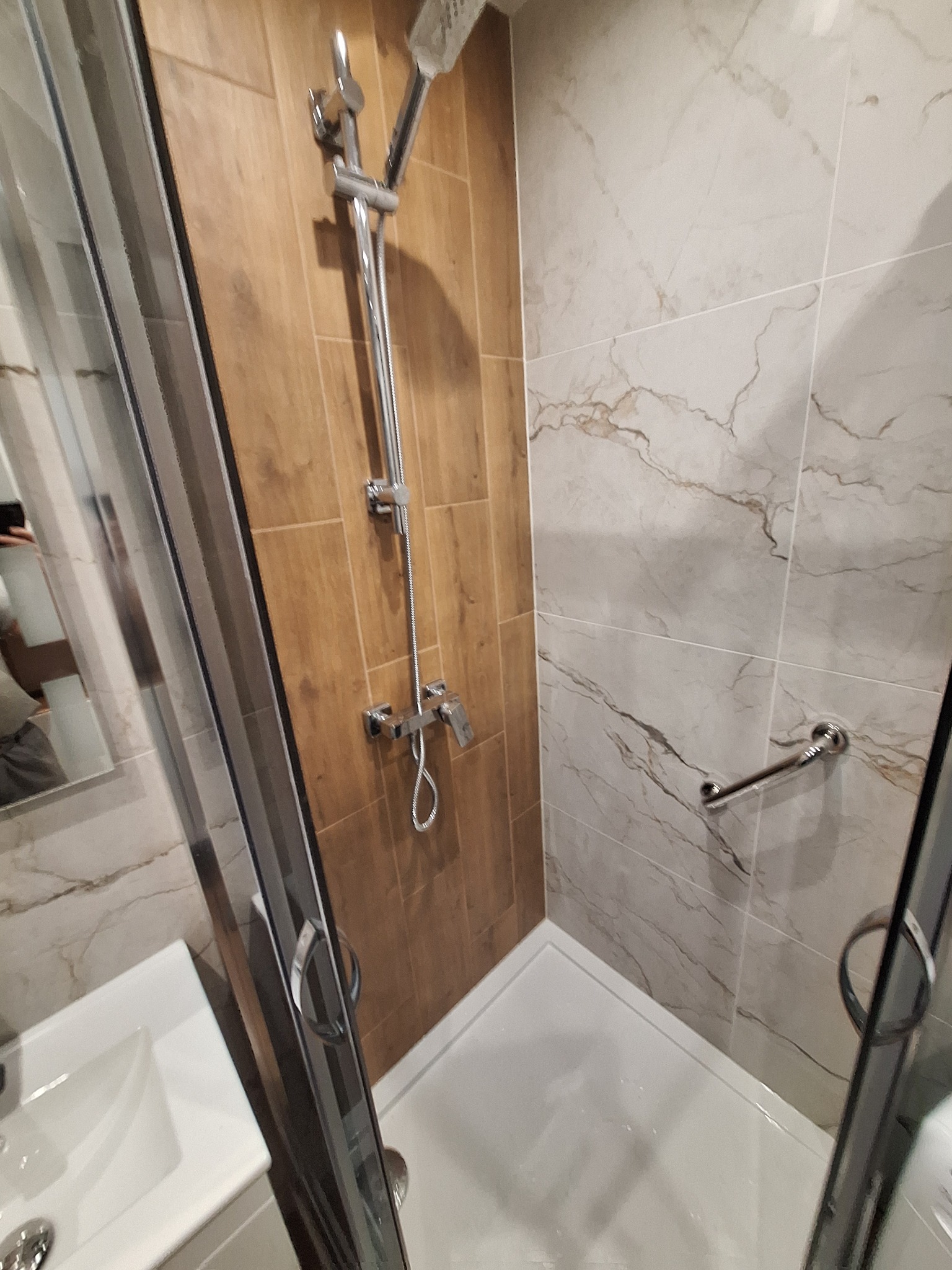 Nowoczesna kabina prysznicowa z chromowaną armaturą, brązowymi i marmurkowymi płytkami oraz uchwytem dla osób z niepełnosprawnościami.